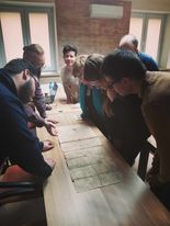 Uczestnicy zebrania pochylają się nad blatem. Na blacie dokumenty archiwalne.