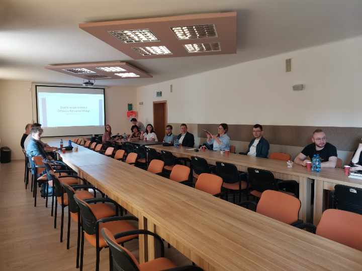 Uczestnicy zebrania siedzący przy stolikach ustawionych w podkowę. W tle rzutnik.