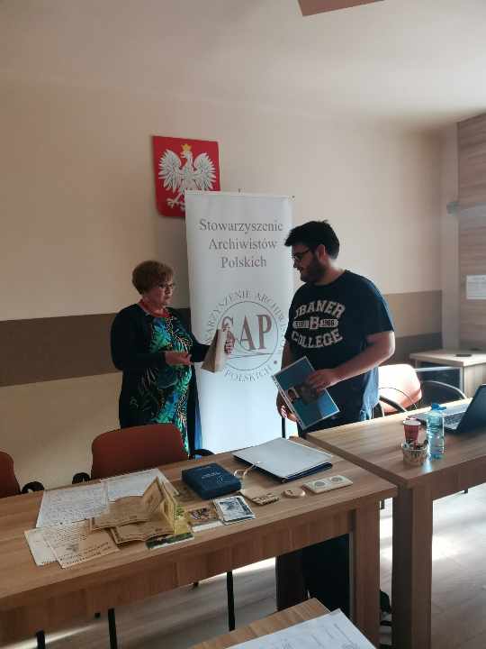 Prezes Julia Dziwoki wręcza Krzysztofowi Kurkowskiemu upominek i plakat. Za nimi baner SAP. Przed nimi stolik z materiałami archiwalnymi.