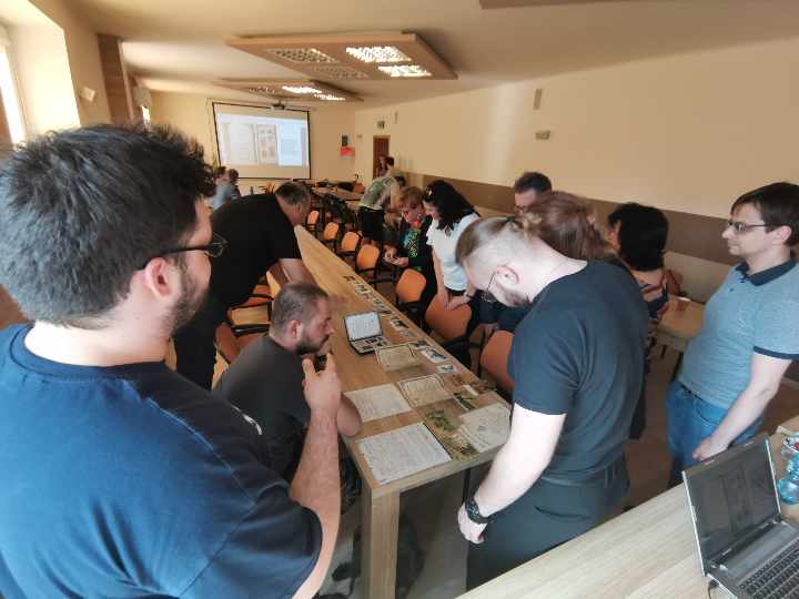 Uczestnicy zebrania stoją wokół stolika, oglądają rozłożone na nim materiały archiwalne.