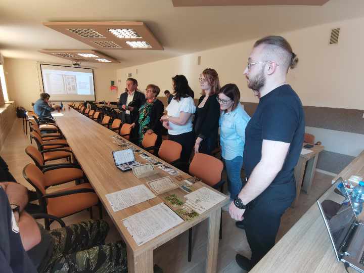 Uczestnicy zebrania stoją wokół stolika, oglądają rozłożone na nim materiały archiwalne.