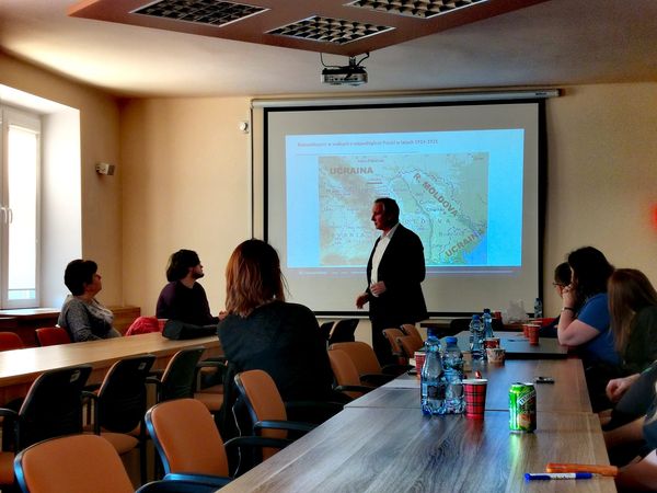 Prelegent na tle mapy Bukowiny. Przed nim uczestnicy zebrania siedzący przy stolikach.