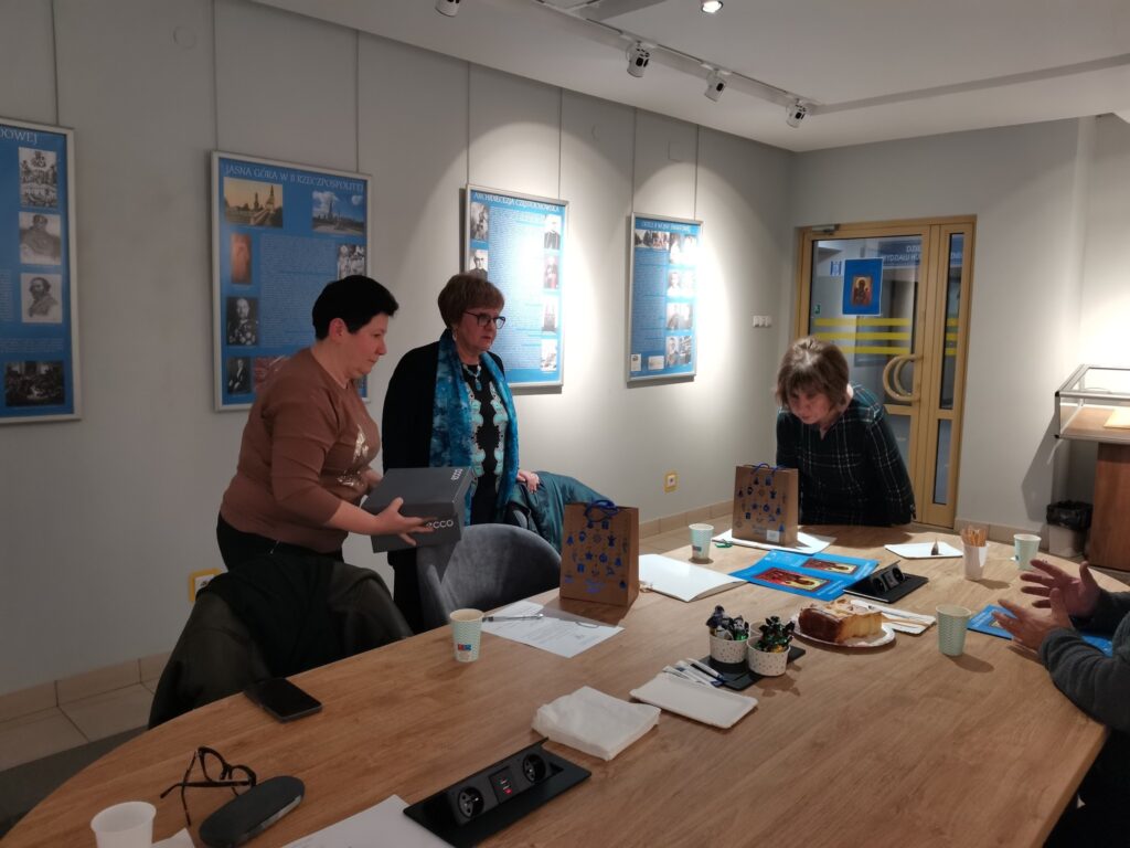 Julia Dziwoki, Beata Urbanowicz i Dorota Wojciechowska na tle wystawy stoją przy stoliku, na którym znajduje się poczęstunek i upominki gwiazdkowe.