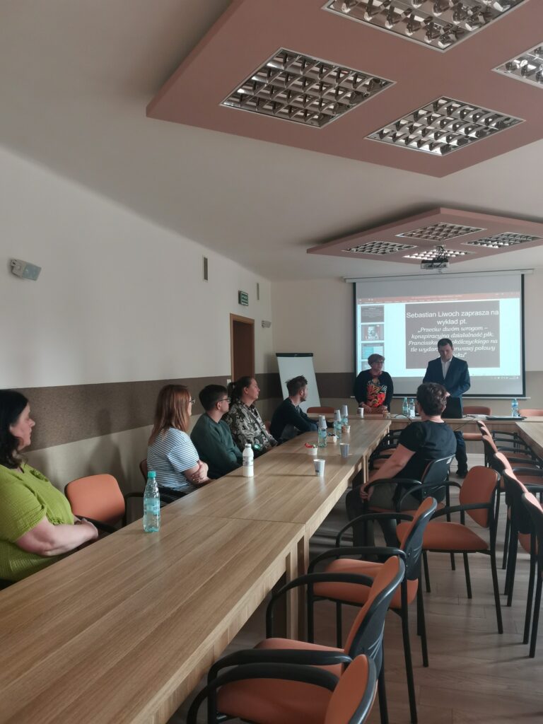 Prezes Julia Dziwoki i prelegent Sebastian Liwoch na tle prezentacji multimedialnej wyświetlanej na rzutniku oraz inni uczestnicy spotkania.