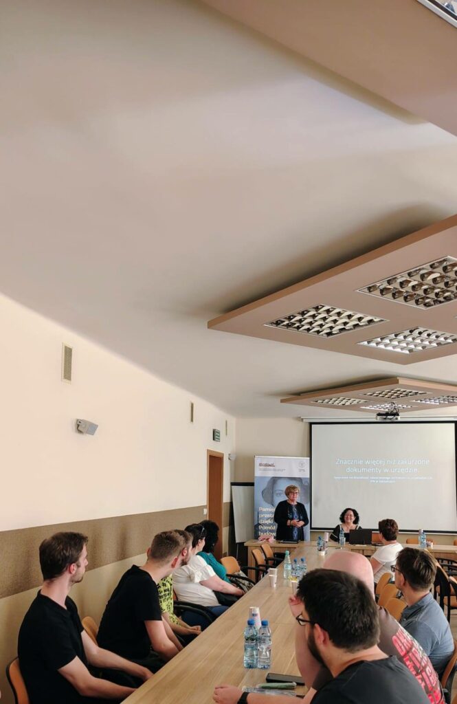 Prezes Julia Dziwoki i prelegentka Renata Dziechcierz na tle prezentacji multimedialnej oraz inni uczestnicy spotkania.