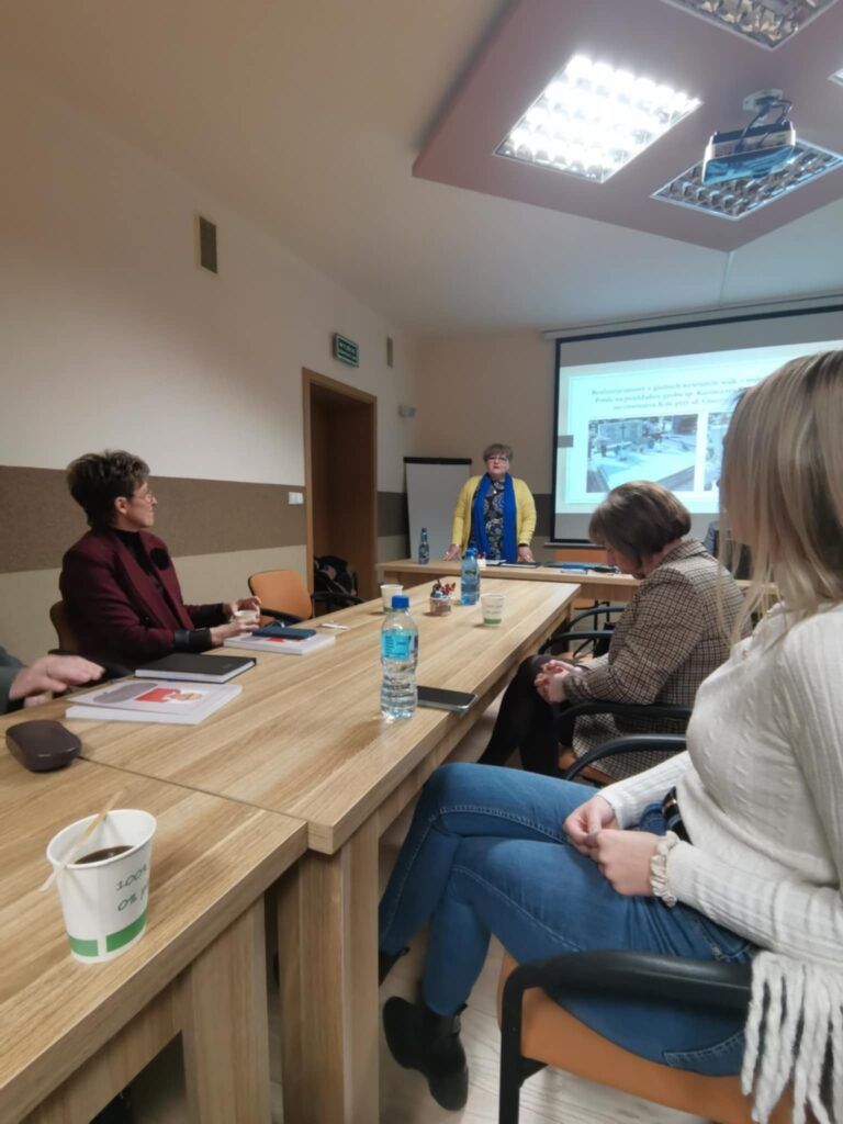 Prezes Julia Dziwoki i uczestnicy zebrania siedzący przy stolikach. W tle prezentacja multimedialna wyświetlona na rzutniku.