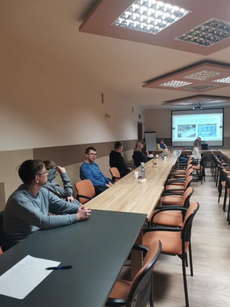 Prezes Julia Dziwoki i uczestnicy zebrania siedzący przy stolikach. W tle prezentacja multimedialna wyświetlona na rzutniku.