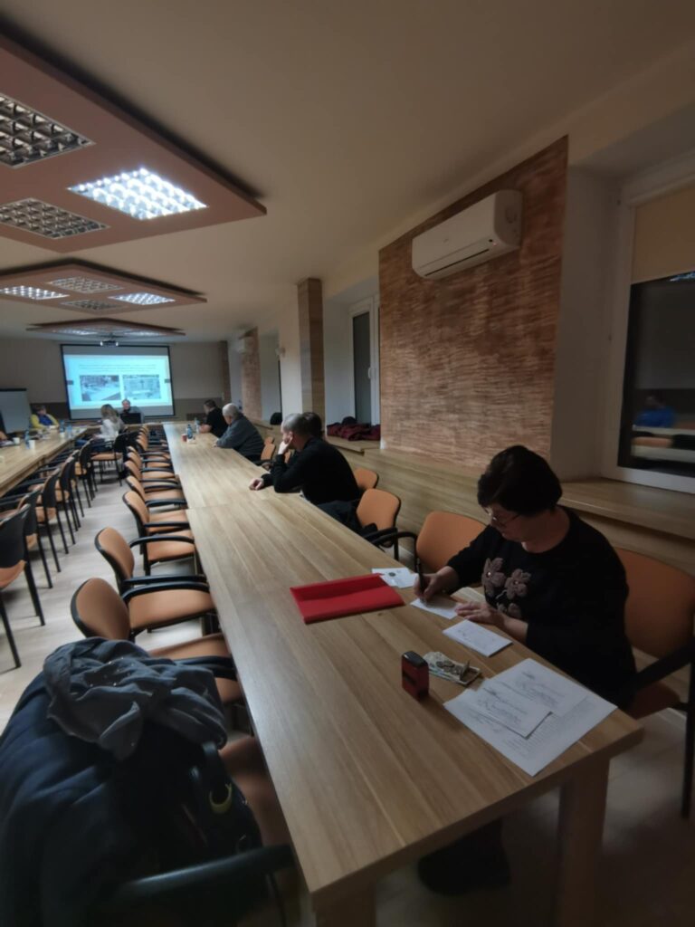 Uczestnicy zebrania przy stolikach ustawionych w podkowę. W tle prezes Julia Dziwoki i prezentacja multimedialna wyświetlona na rzutniku.