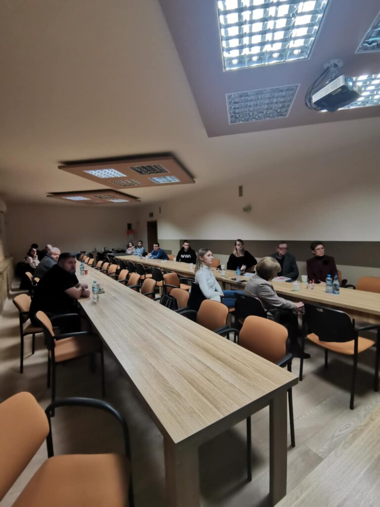 Uczestnicy zebrania przy stolikach ustawionych w podkowę. 