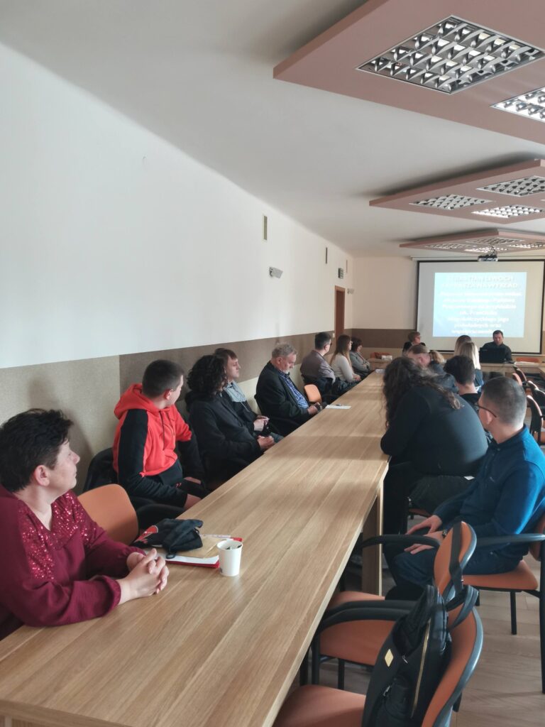 Uczestnicy spotkania przy stolikach ustawionych w podkowę. W tle prelegent - Sebastian Liwoch.