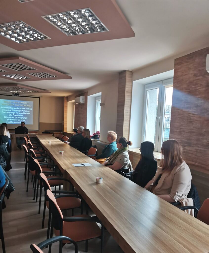 Uczestnicy spotkania przy stolikach ustawionych w podkowę. W tle prelegent - Sebastian Liwoch.